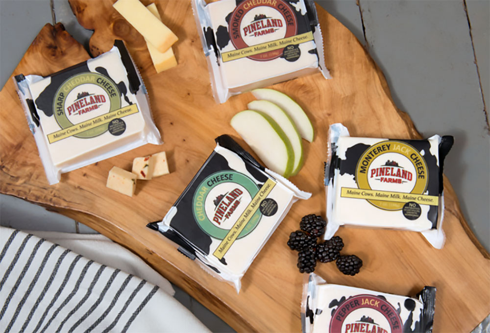 Multiple blocks of cheese on a butcher block table with scattered grapes, and fruit slices and a Pineland Farms Dairy sticker.